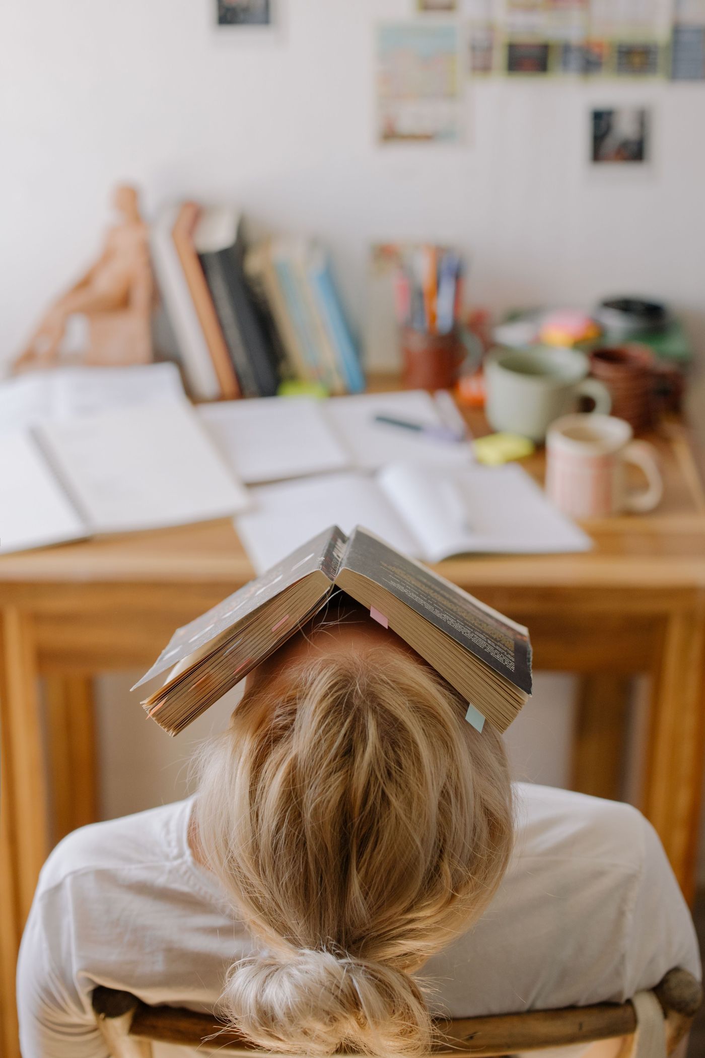 A student slumped at a desk with an open book resting on their head, study books and mugs behind