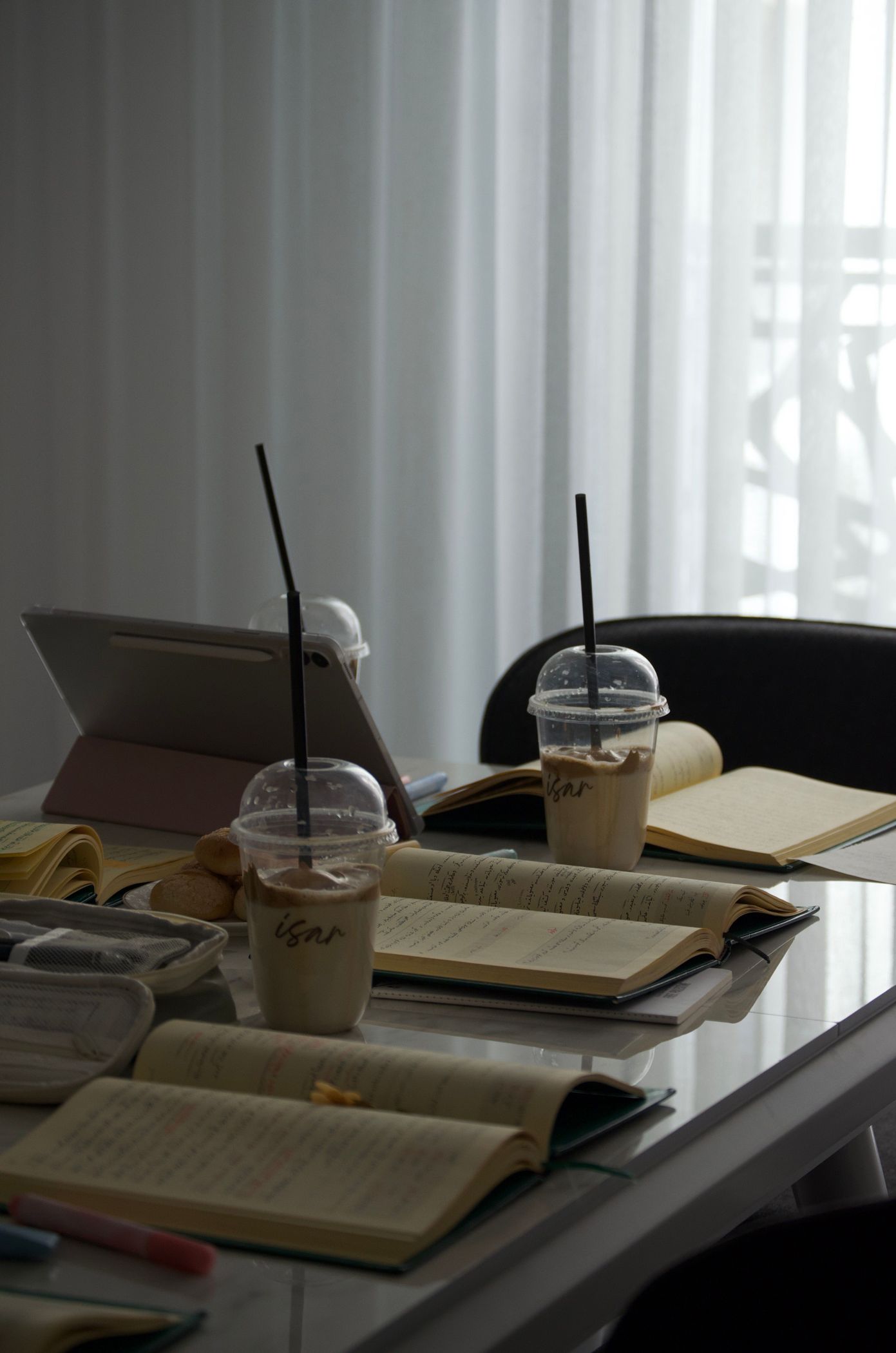 A study table with open notebooks, a tablet and two iced coffees by a curtained window