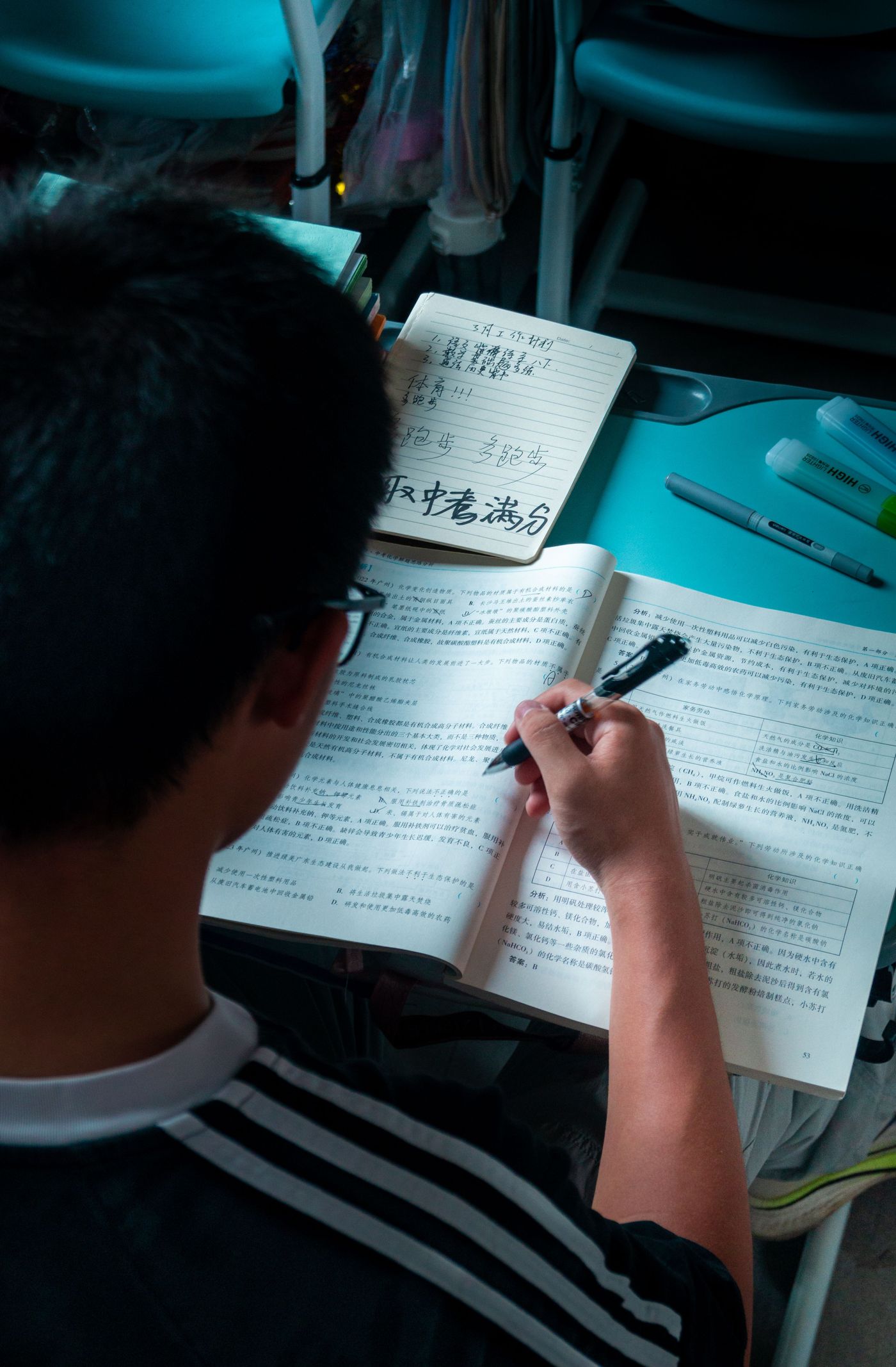 A student writing in an open test-prep workbook, a notebook of handwritten notes propped above it