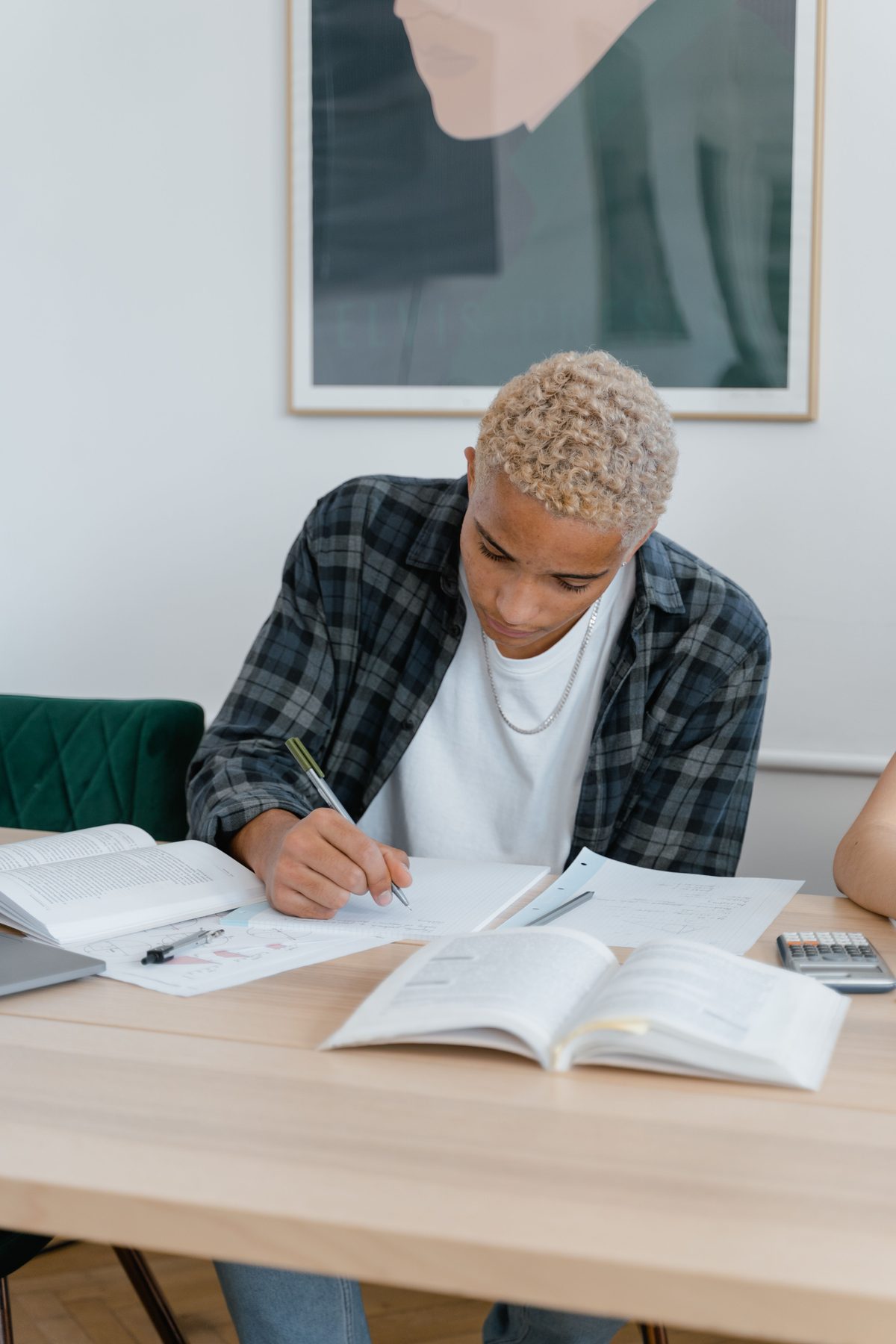 A student in a plaid shirt writing notes at a wooden table, surrounded by open study books