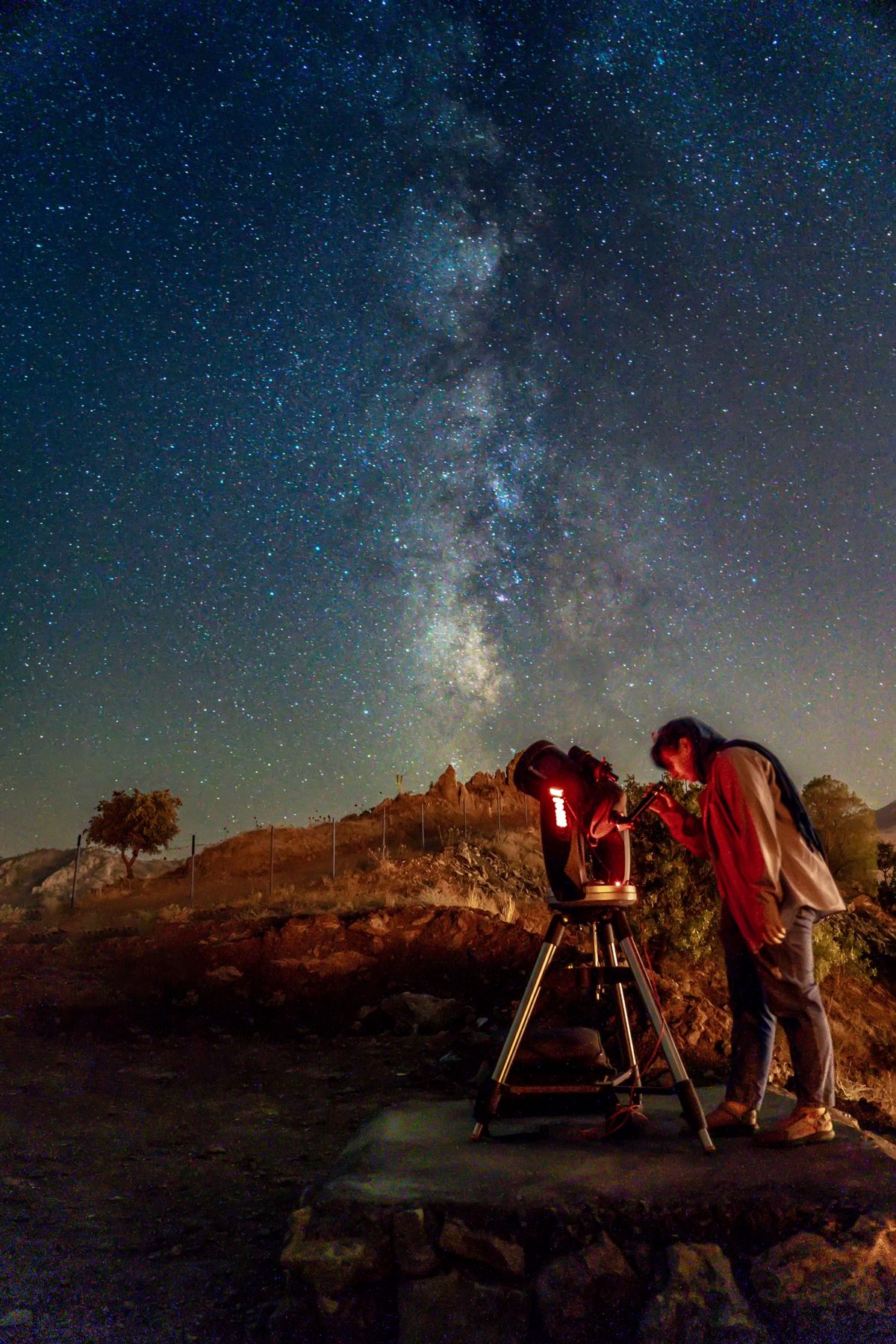 A person looking through a telescope on a hillside at night, under the Milky Way