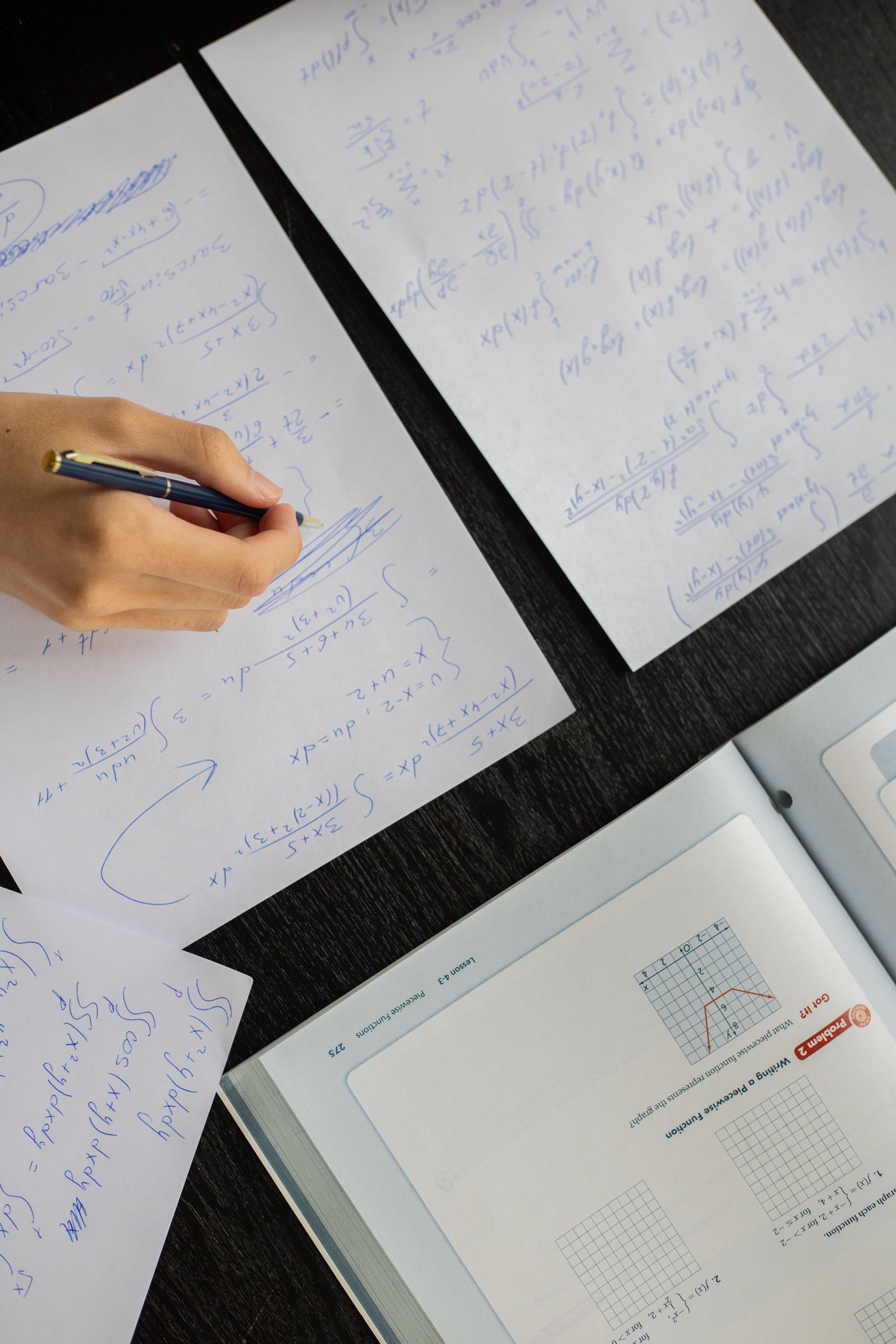 A hand writing out calculus working in blue ink on sheets of paper beside an open maths textbook