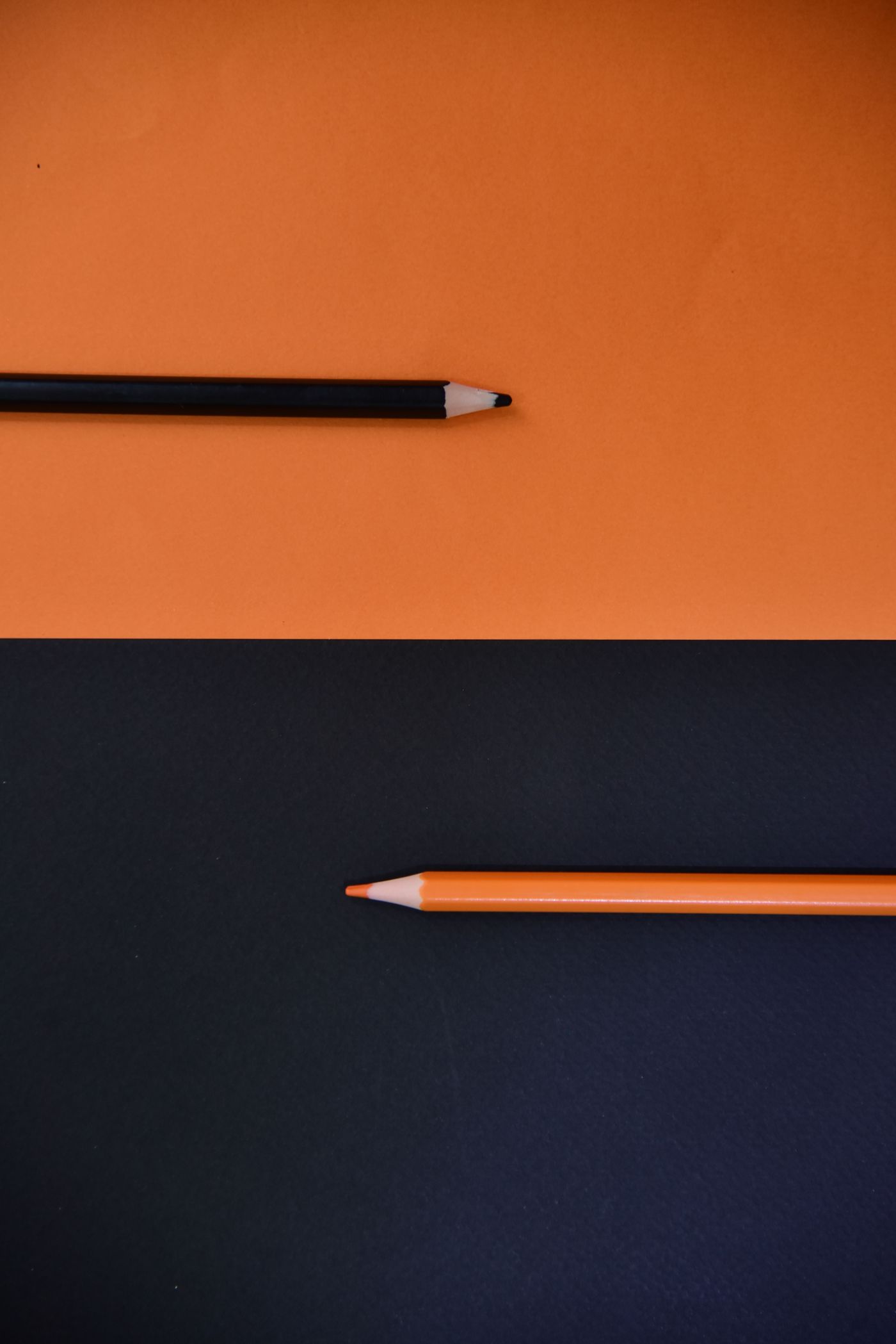 A black pencil on orange paper and an orange pencil on black paper, pointing towards each other