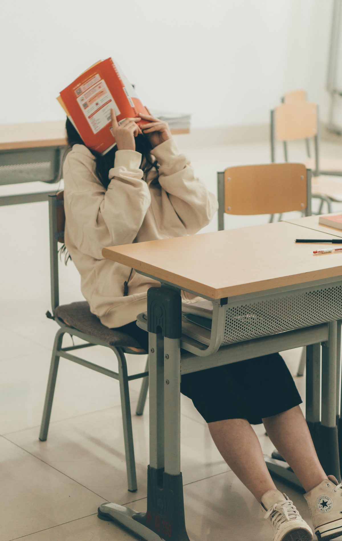 A student at a classroom desk hiding her face behind an open orange textbook