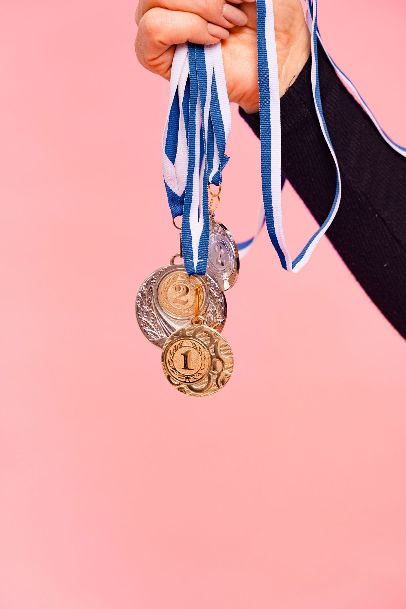 A hand holding gold, silver and bronze medals on blue ribbons against a pink background
