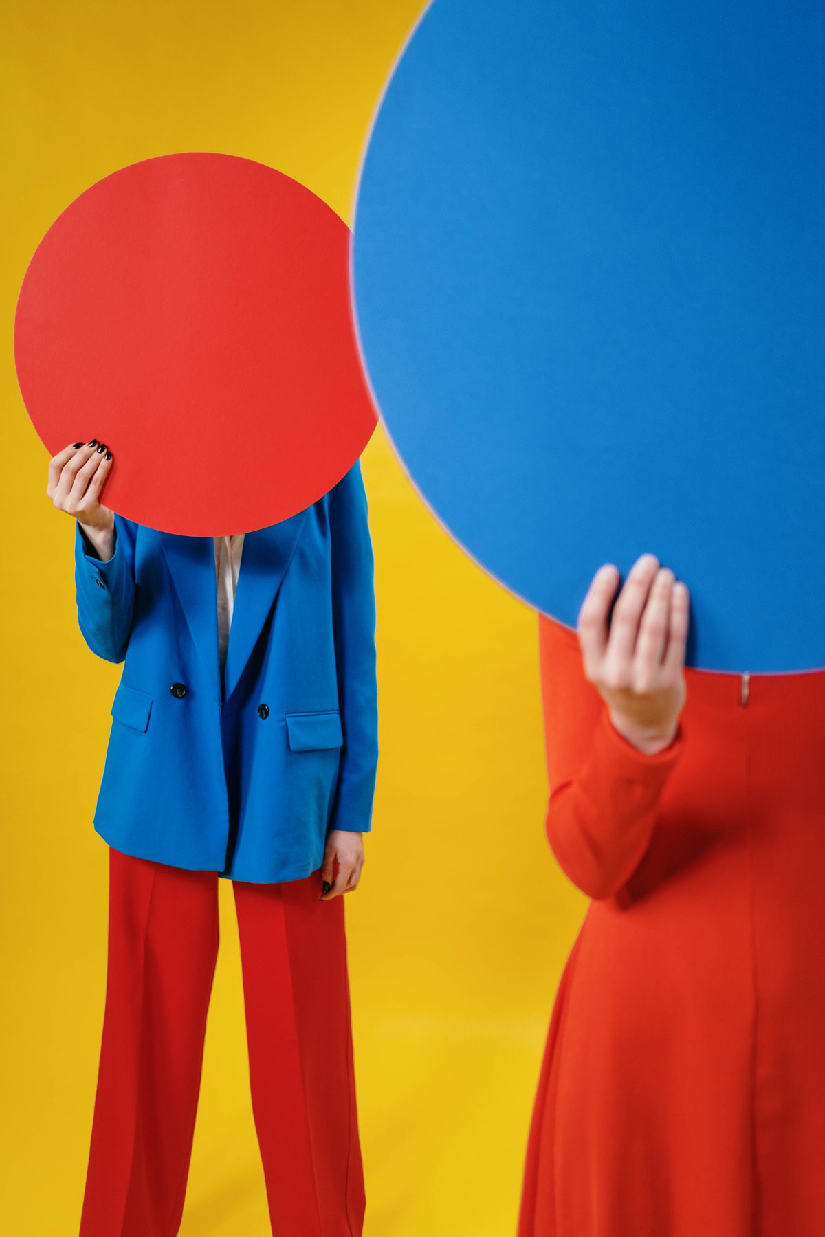 Two people in bright blue and red outfits, each holding a large flat circle in front of their face, against a yellow background.