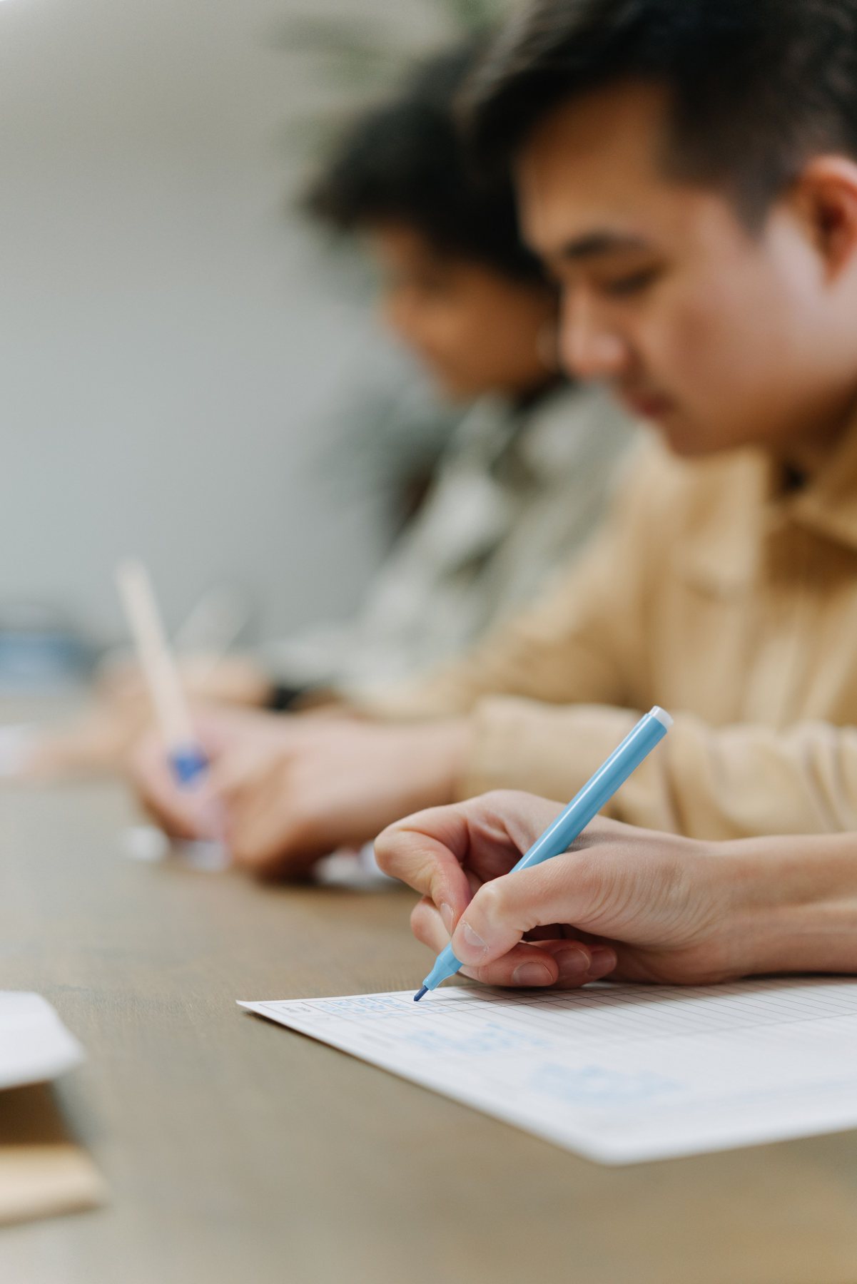 Two students writing on answer sheets at a desk, one blue pen in sharp focus in the foreground.