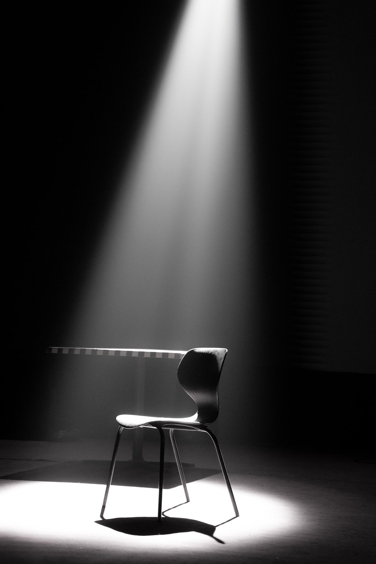 A single empty chair and table lit by a dramatic beam of light in a dark room.
