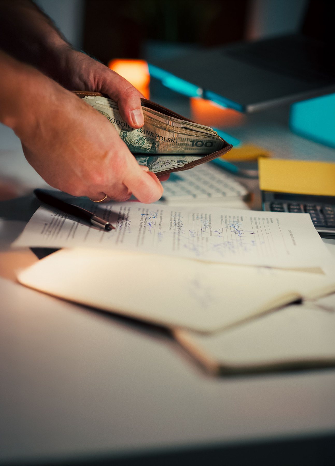 A hand holding a wallet of banknotes over a desk covered with papers, a pen, notebooks and a calculator.