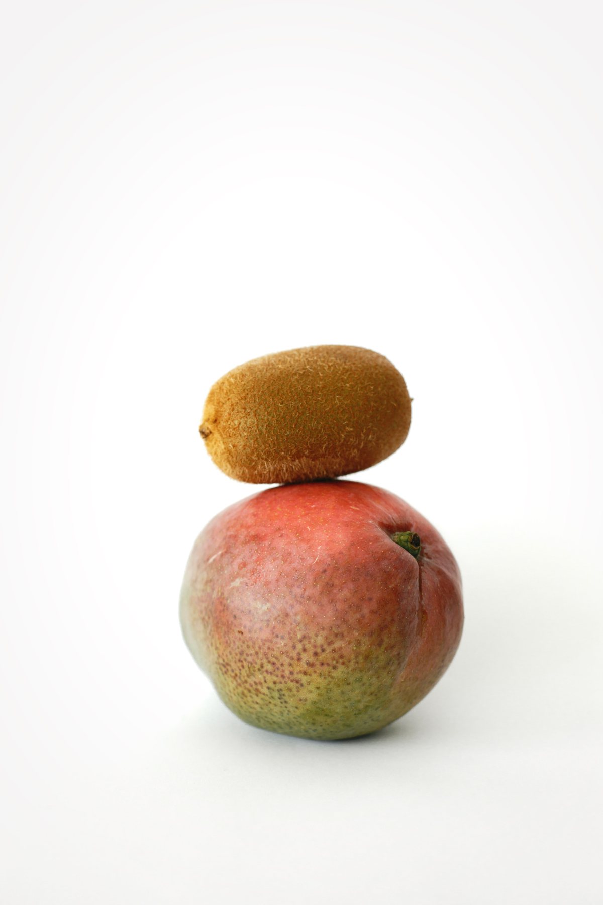 Two pieces of fruit, a small kiwi balanced on top of a larger mango, against a plain white background.