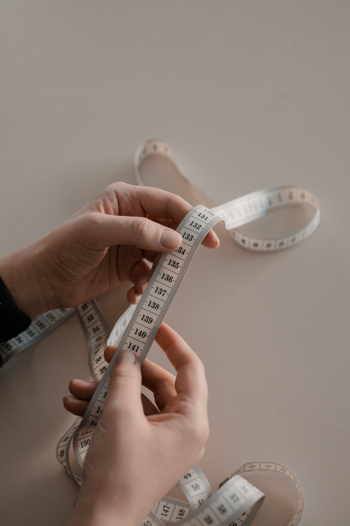 Two hands holding a cloth measuring tape stretched between them on a pale table.