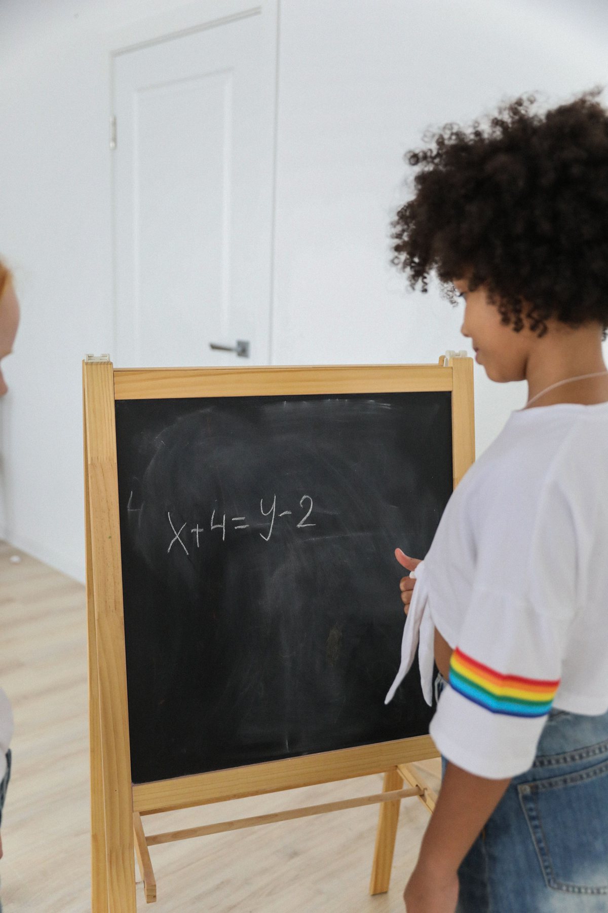 A child in a white t-shirt stands at a wooden chalkboard easel with the equation x + 4 = y - 2 written on it.